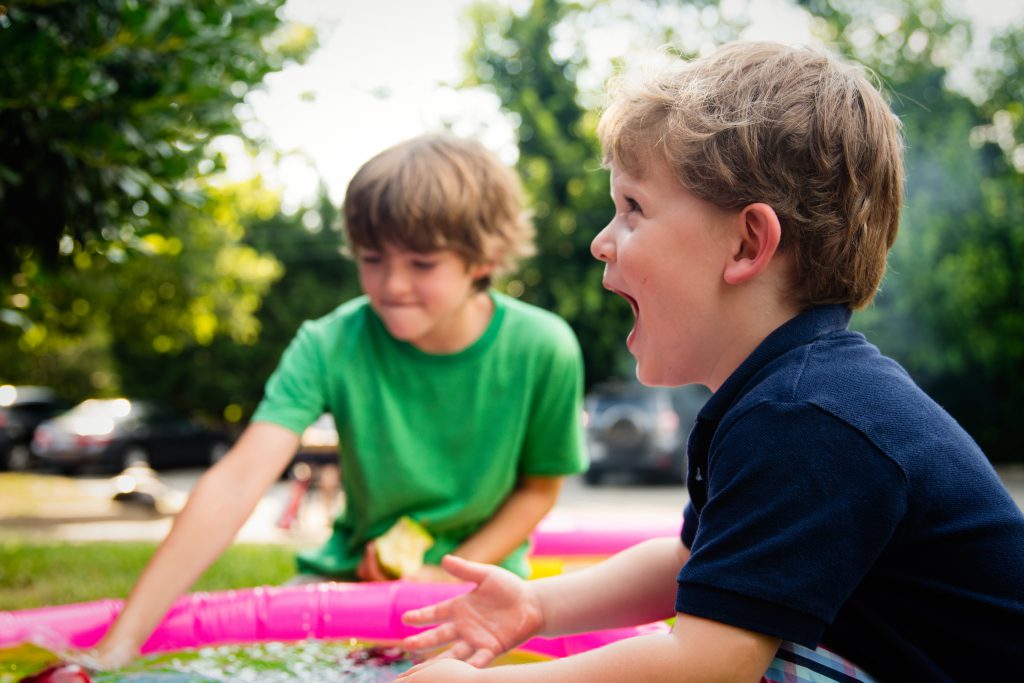 Zwei Kinder spielen im Garten mit einem Gummischlauch. Das Kind vorne im Bild schreit. 
