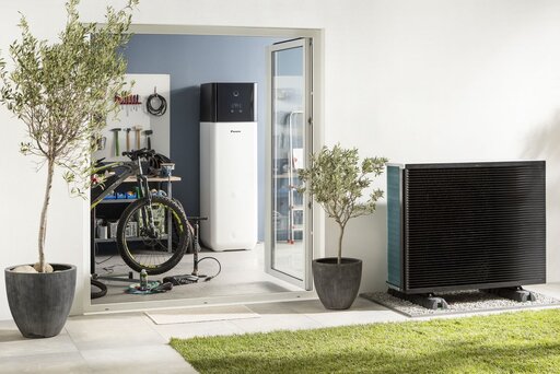 Eine moderne Immobilie in Anthrazit und Weiß mit Terrasse und Garten. Auf dem Rasen steht eine zylindrische Wärmepumpe in silber-anthrazitfarbener Optik, die wie ein Kunstwerk wirkt.