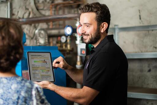 ALT-Text: Junger Mann mit Bart und dunklen Haaren in Polohemd zeigt einer Dame, die man nur von hinten sieht, auf einem Tablet den Energieausweis. 