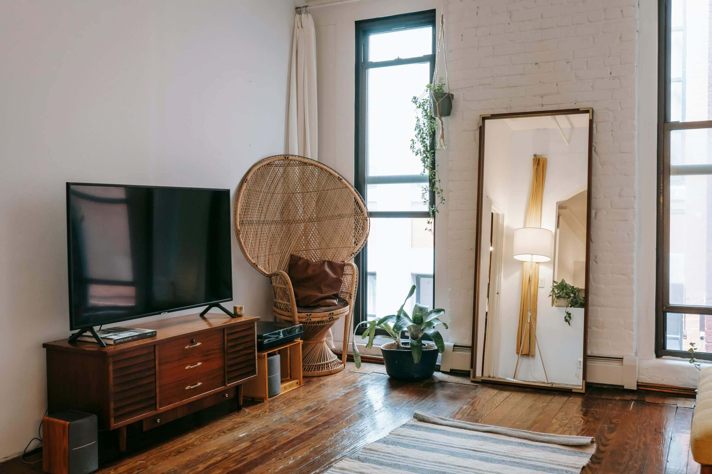 Gemütliches Wohnzimmer mit einem großen Spiegel, einem Rattansessel, Pflanzen und einem Holz-Fernsehschrank mit Smart TV.