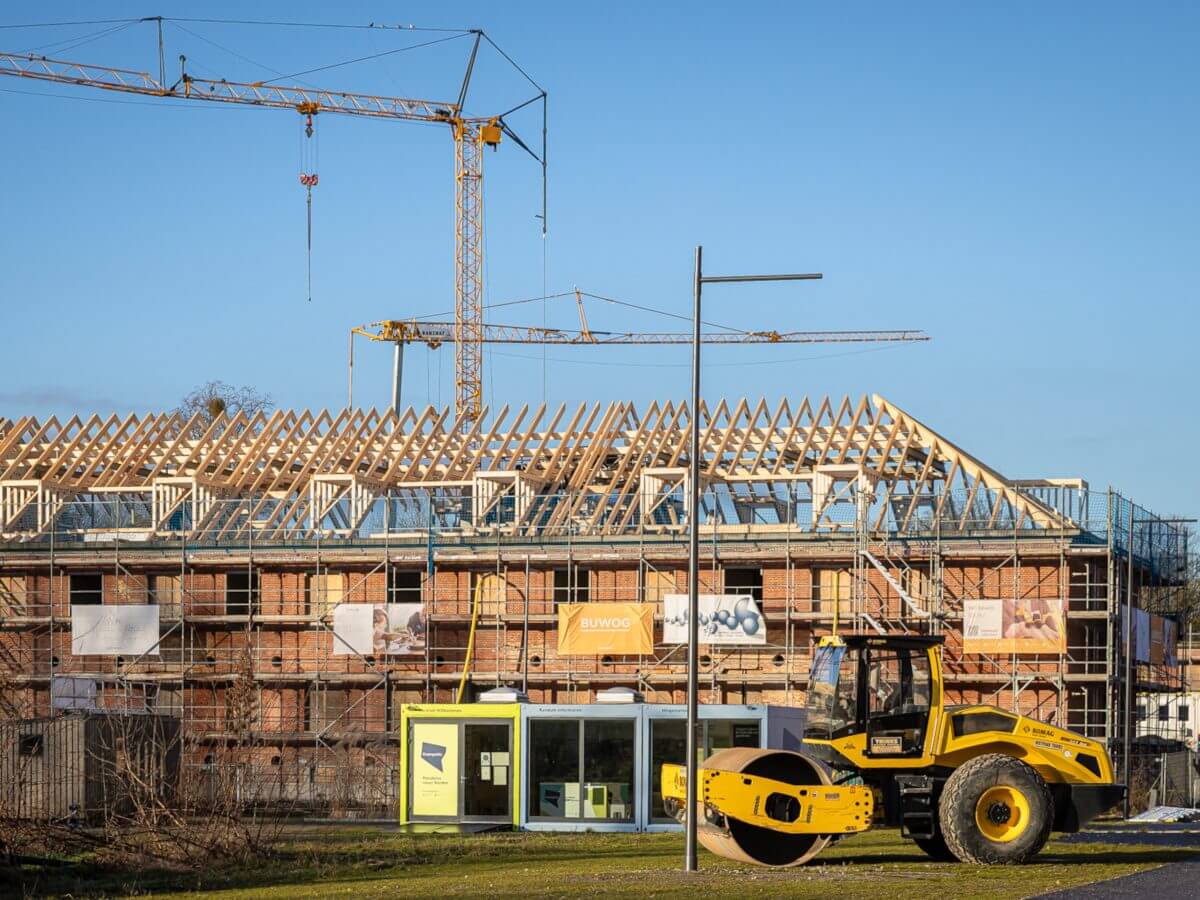 Wohngebäude von BUWOG im Bau mit Holz-Dachkonstruktion, zwei Kränen und einer gelben Straßenwalze im Vordergrund.