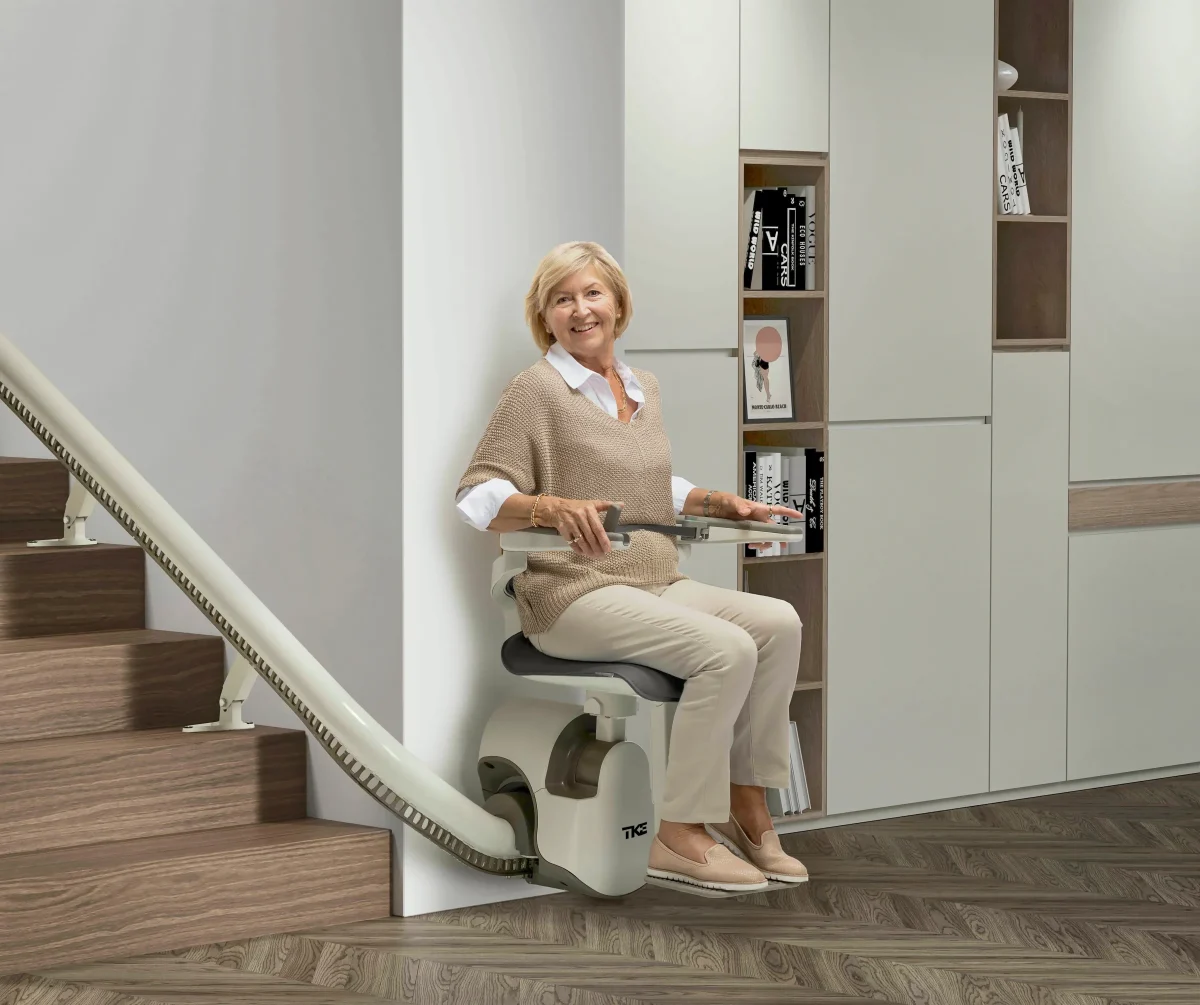 Eine Frau mittleren Alters mit blonden Haaren sitzt auf einem Treppenlift am unteren Ende der Treppe. Ihr Blick ist in die Kamera gerichtet.