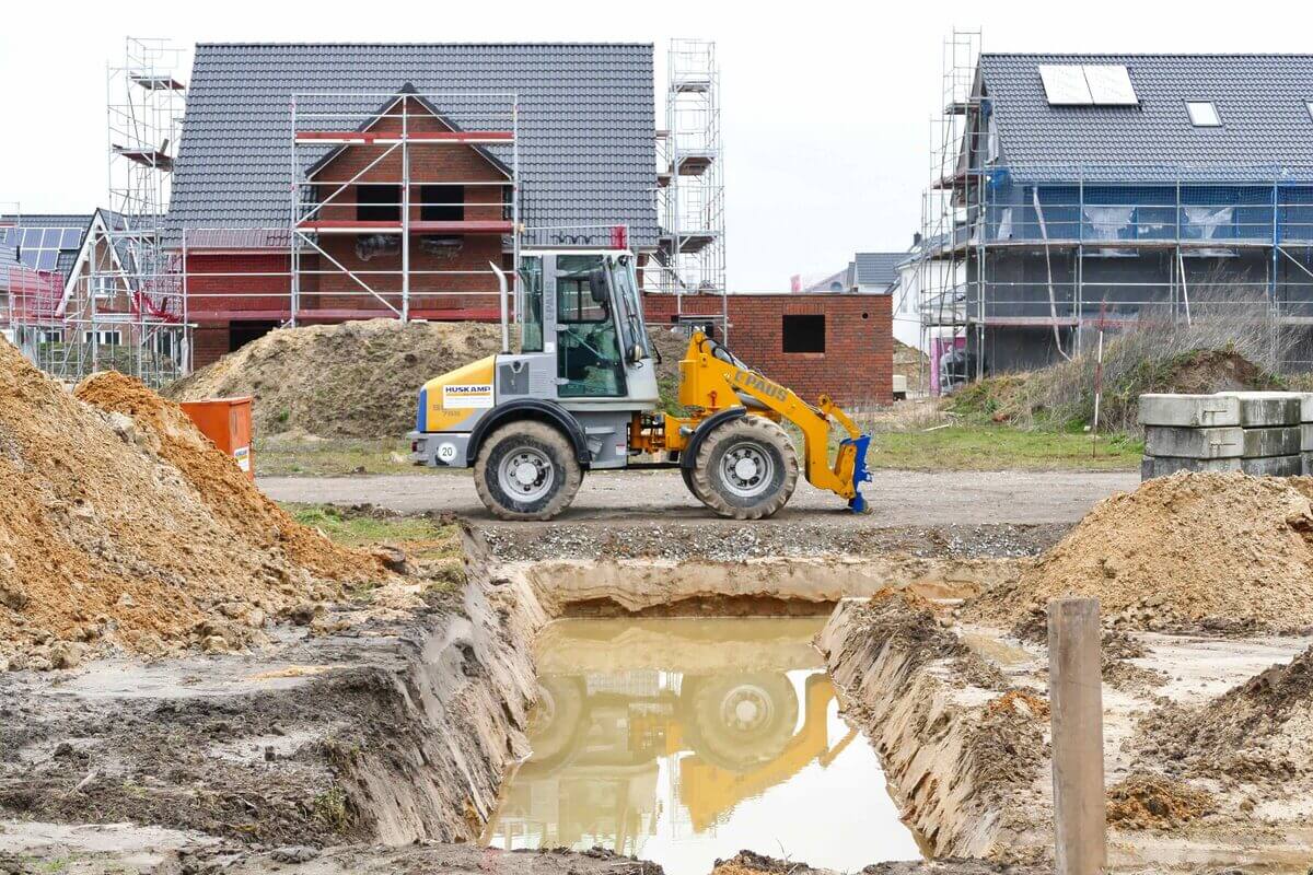 Ein Baufahrzeug fährt in einem Neubaugebiet vor zwei Neubau-Häusern im Rohbau. 